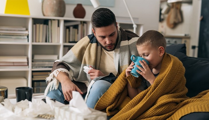 Ein Junge auf einem Sofa ist in eine wärmende Decke eingehüllt und trinkt aus einer Tasse, neben ihm ein Erwachsener