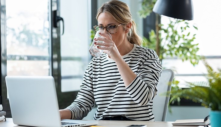 Frau sitzt vor dem Laptop am Schreibtisch und trinkt ein Glas Wasser