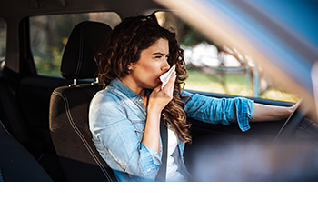 Eine Frau sitze am Steuer eines Autos und hält sich hustend ein Taschentuch vor den Mund