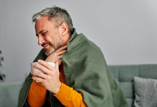 Ein Mann mit einer Teetasse in der Hand sitzt in eine Decke eingehüllt auf dem Sofa und greift sich mit schmerzverzerrtem Gesicht an den Hals