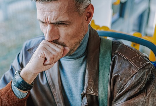 Ein Mann mit grauen Haaren sitzt im Bus, hustet und hält sich dabei die Hand vor den Mund